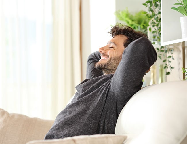 Smiling Caucasian man leaned back on couch with his hands behind his head