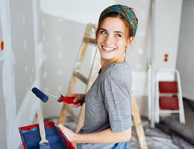 Woman wearing gray shirt and headband smiling while holding paint roller full of blue paint in sheetrocked room