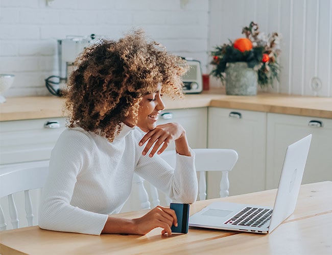 African American woman sitting in kitchen smiling at laptop while holding credit card