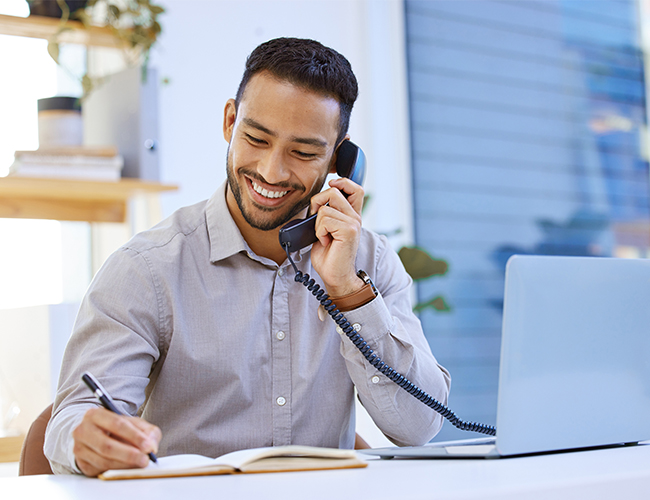 Smiling Asian man talking on phone while writing in notebook in front of laptop Smiling Asian man talking on phone while writing in notebook in front of laptop